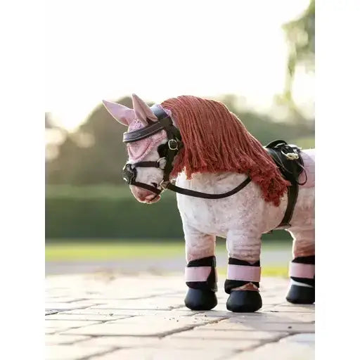 A plush pink lemieux toy pony with a red knitted pony fly hood blossom and black leather harness and hoof covers