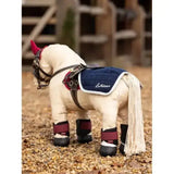 Cream-colored lemieux toy pony wearing a navy quilted toy pony quarter sheet with red ear covers and maroon ankle wraps