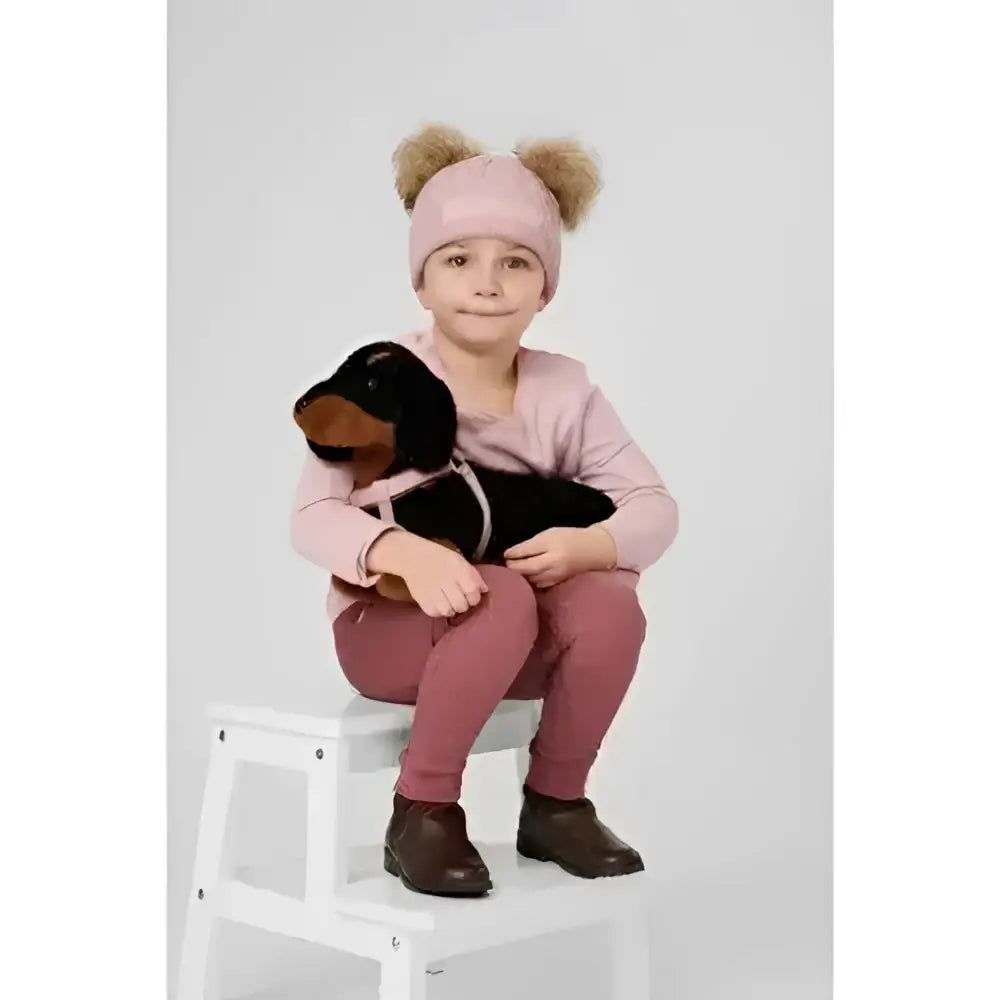 A child in a pink beanie holds a black lemieux toy puppy sally plush dachshund sitting on a white stool