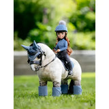 A toy rider olivia doll wearing a blue helmet and outfit sits on a white toy pony with gray leg wraps and black bridle