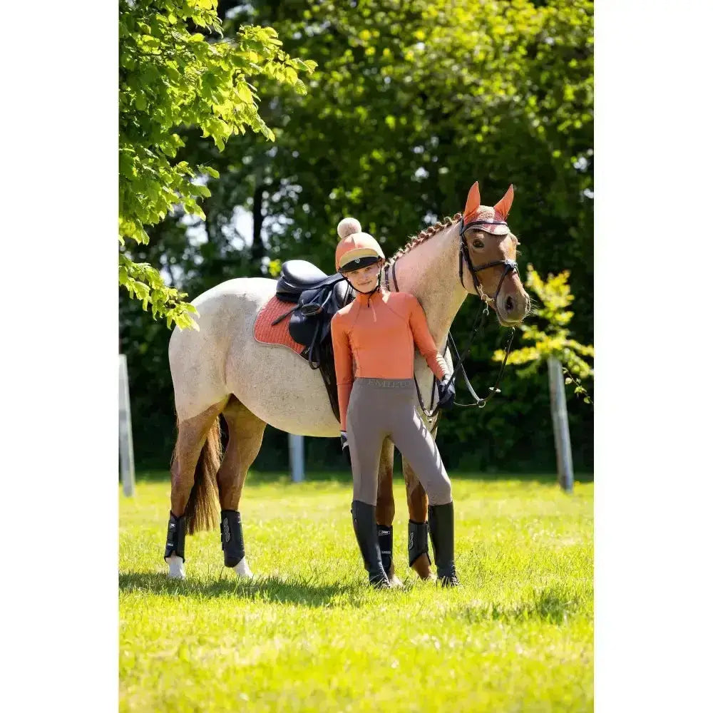 A light-colored horse with dark mane wearing an orange bridle next to a lemieux young rider base layer apricot