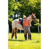 A light-colored horse with dark mane wearing an orange bridle next to a lemieux young rider base layer apricot