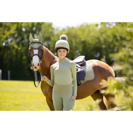 A woman wearing a sage green lemieux young rider base layer fern stands beside a brown horse with black bridle and saddle