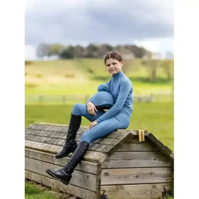 A woman wearing a lemieux young rider base layer ice in blue sits on a wooden structure with black knee-high boots