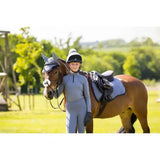 A young rider in a grey lemieux young rider base layer jay blue stands beside a brown horse with silver ear protection