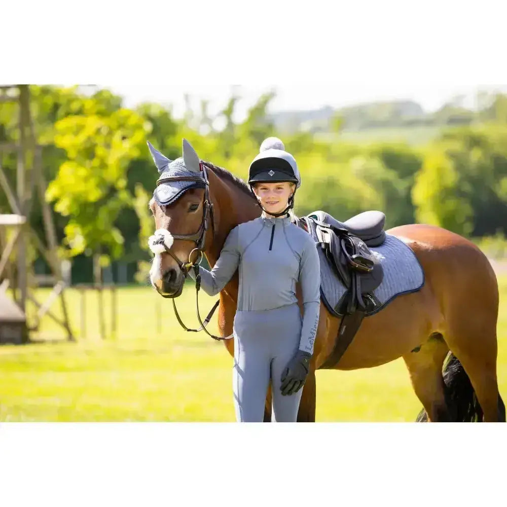 A young woman wears a light gray lemieux young rider base layer jay blue standing next to a brown horse