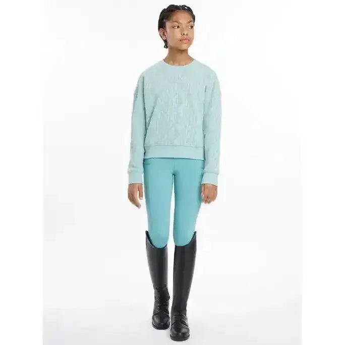 A young girl wearing lemieux young rider clea crew sweat aqua long-sleeved sweater paired with turquoise riding pants