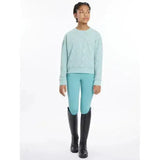 A young girl wearing lemieux young rider clea crew sweat aqua long-sleeved sweater paired with turquoise riding pants