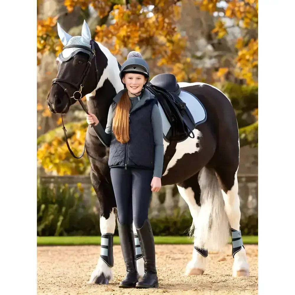 Black and white pinto horse wearing silver ear covers beside lemieux young rider eloise reversible winter gilet navy