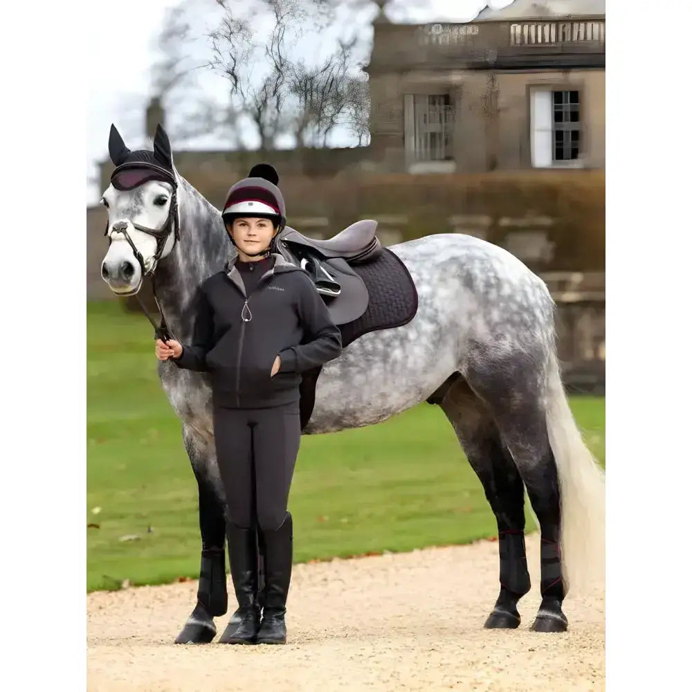 A young rider in a black lemieux young rider hollie lined hoodie stands beside a dapple grey horse with dark tack