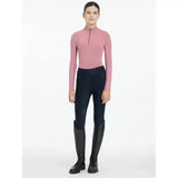 A woman wears the lemieux young rider hope lightweight base layer peony pink equestrian top with black riding pants and boots