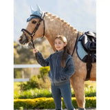 A young girl wearing lemieux young rider libby fleece petrol navy blue fleece stands next to a horse with matching blue tack