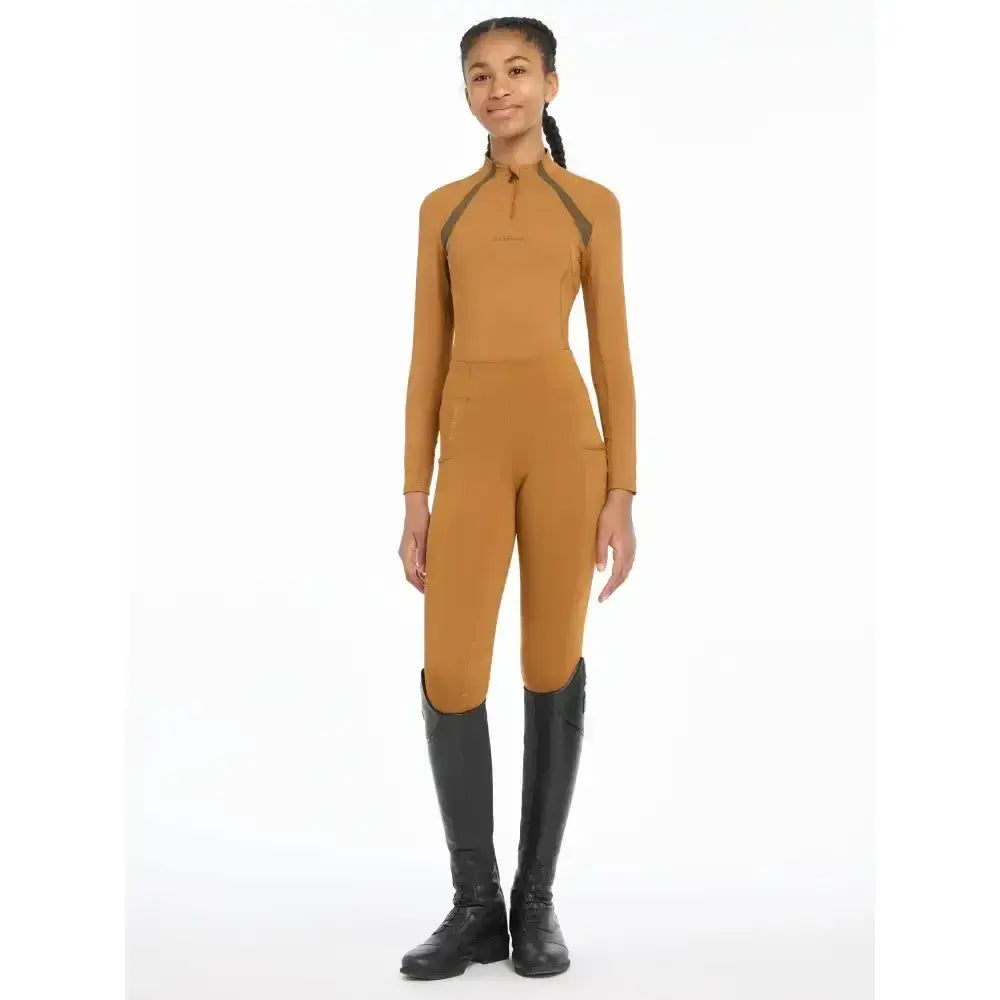 A girl wearing a tan lemieux young rider mia mesh base layer jumpsuit with dark gray panels on shoulders and sides