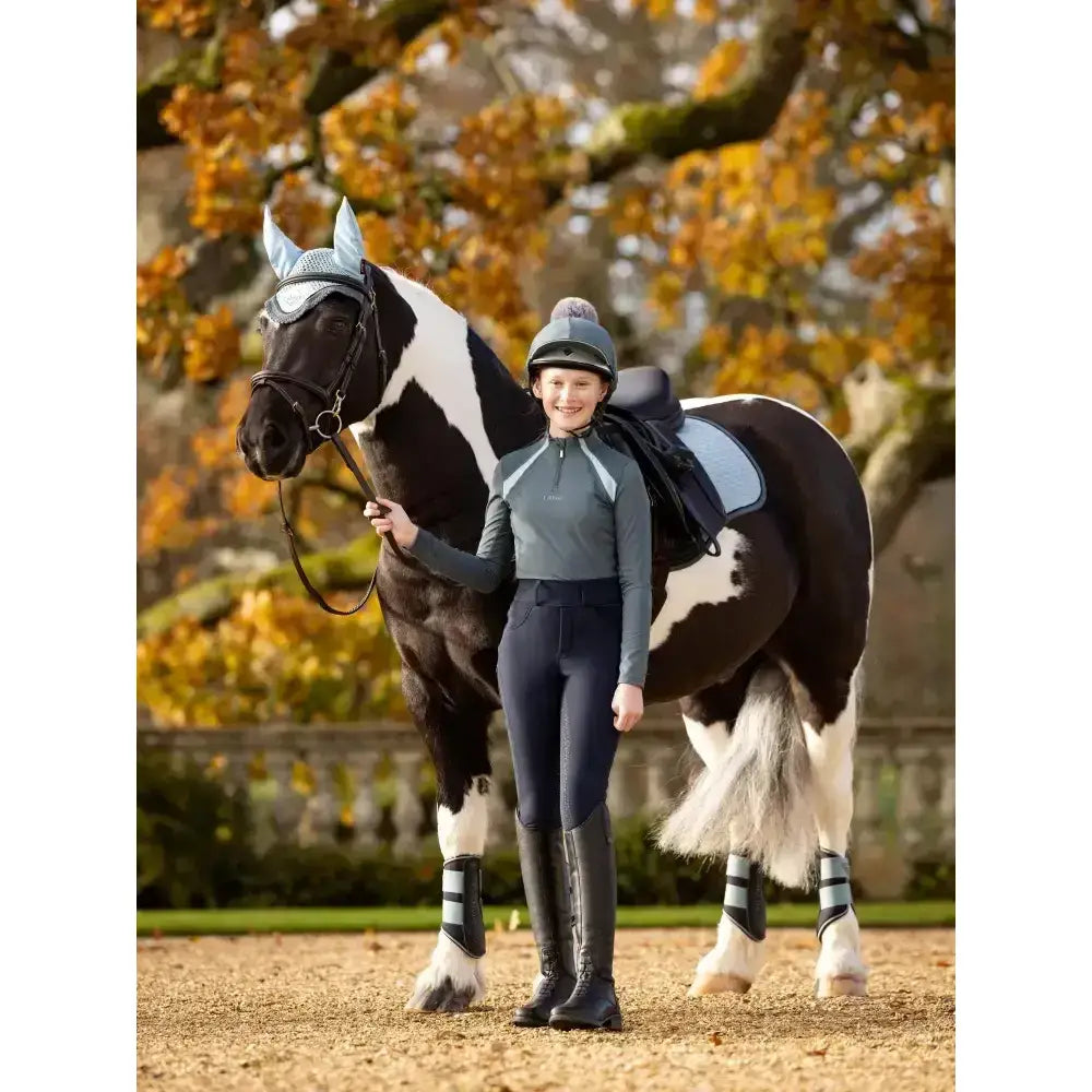 Black and white pinto horse wearing silver ear covers featuring lemieux young rider mia mesh base layer petrol