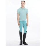 A woman wearing a lemieux young rider polo shirt in aqua paired with turquoise riding pants and black boots