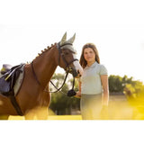 Brown horse with white blaze wearing black bridle next to lemieux young rider polo shirt pistachio in dark saddle pad