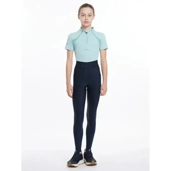 A woman wears lemieux young rider remi breggings navy with black riding pants and a light blue short sleeved polo shirt