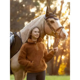 A young girl wearing a rust-colored lemieux young rider tia teddy fleece stands beside a cream horse with bridle