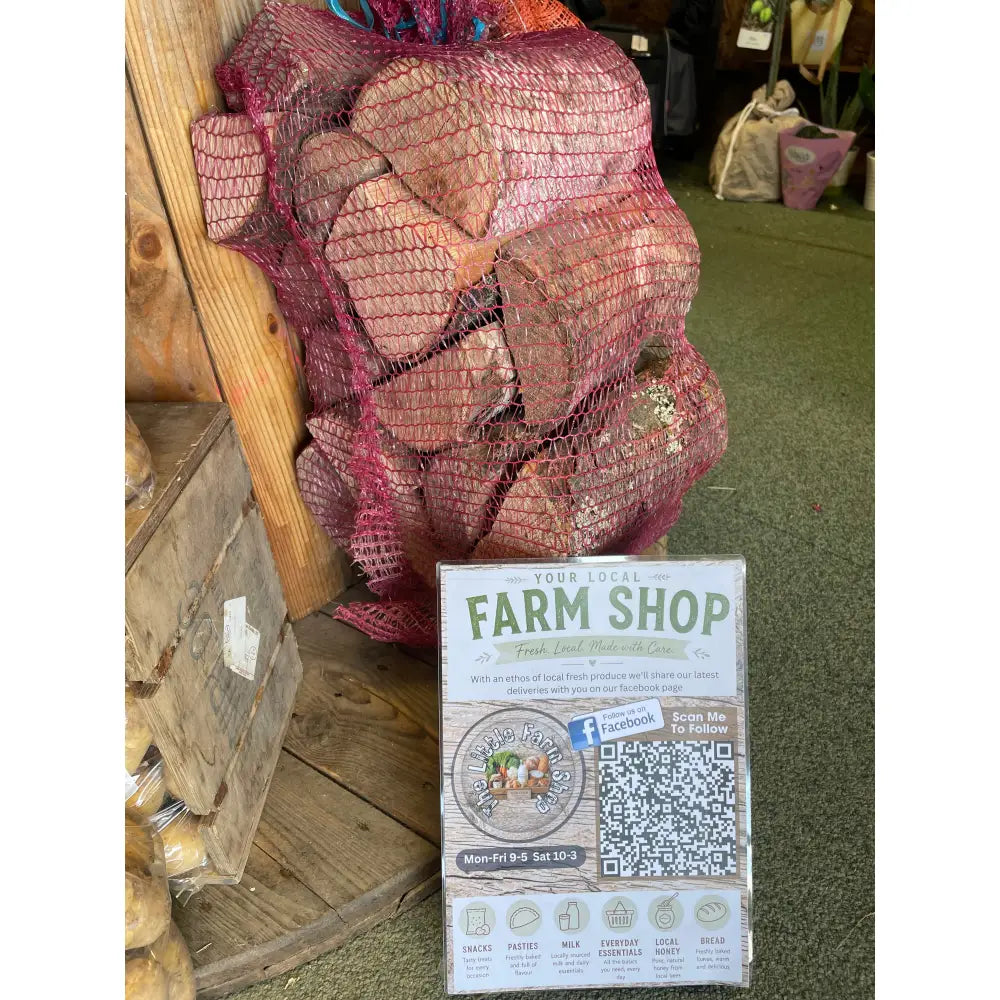 A red mesh bag of heart-shaped potatoes sits beside a wooden crate and farm shop sign near locally cut hardwood logs