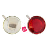 A pink miles tea berry envelope teabags tag next to a clear glass cup of vibrant red coffee berry brewed tea