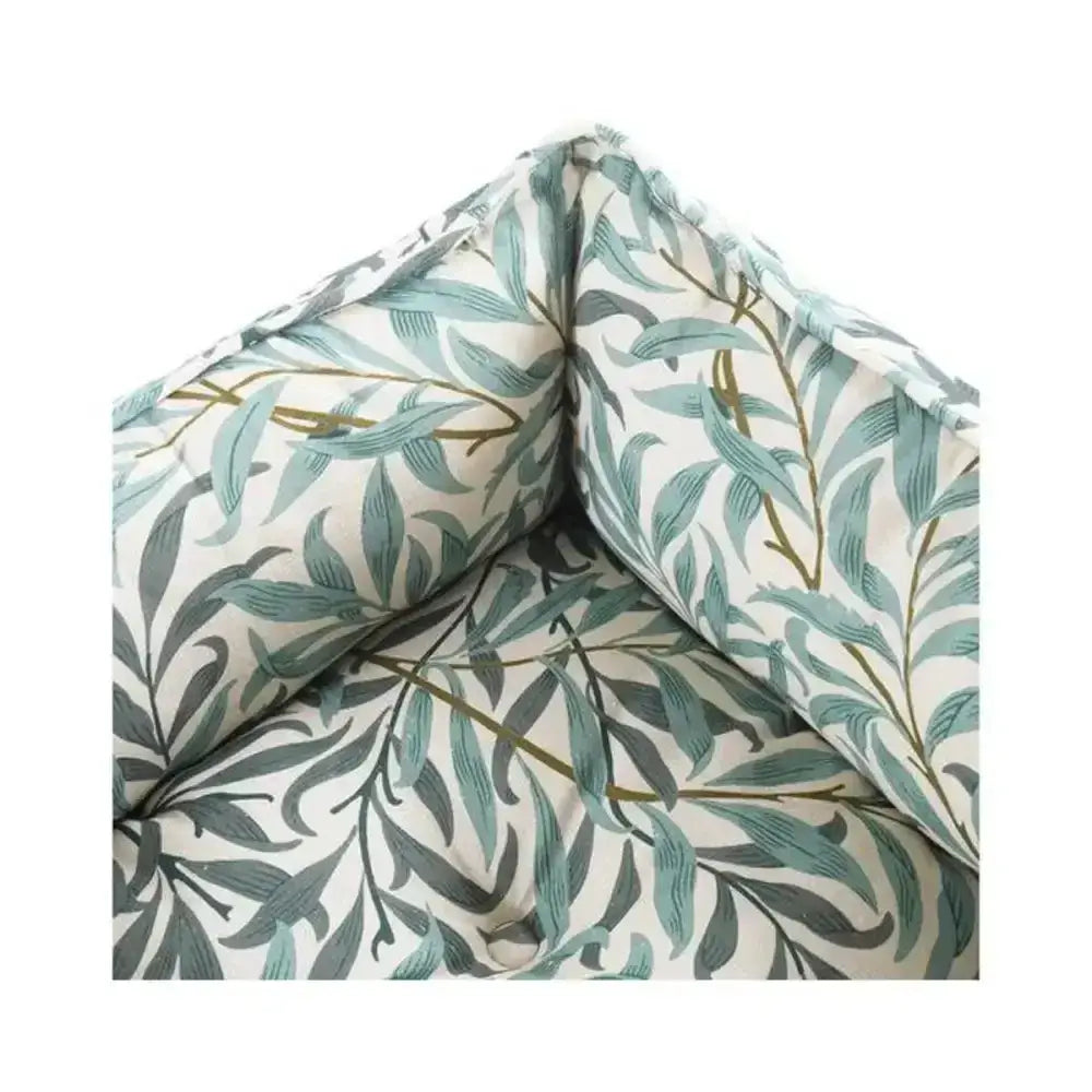 A soft cushioned corner protector in cream fabric with teal and dark green leaves on a morris and co willow boughs print bed