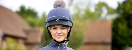 Navy blue equestrian helmet with a fuzzy gray pom-pom on top and a small diamond-shaped logo on the front.