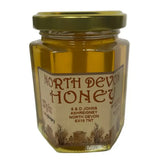 A hexagonal glass jar of golden raw north devon honey sealed with a gold metal lid labeled north devon honey