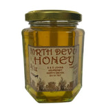 A hexagonal glass jar of golden raw spring blossom north devon honey sealed with a gold metal lid and s & d johns label