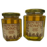 Two hexagonal glass jars of golden north devon honey with gold lids labeled s and d johns raw spring blossom honey