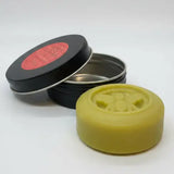 A round olive green beeswax candle with a bee design next to an open black tin for north devon honey cream bar