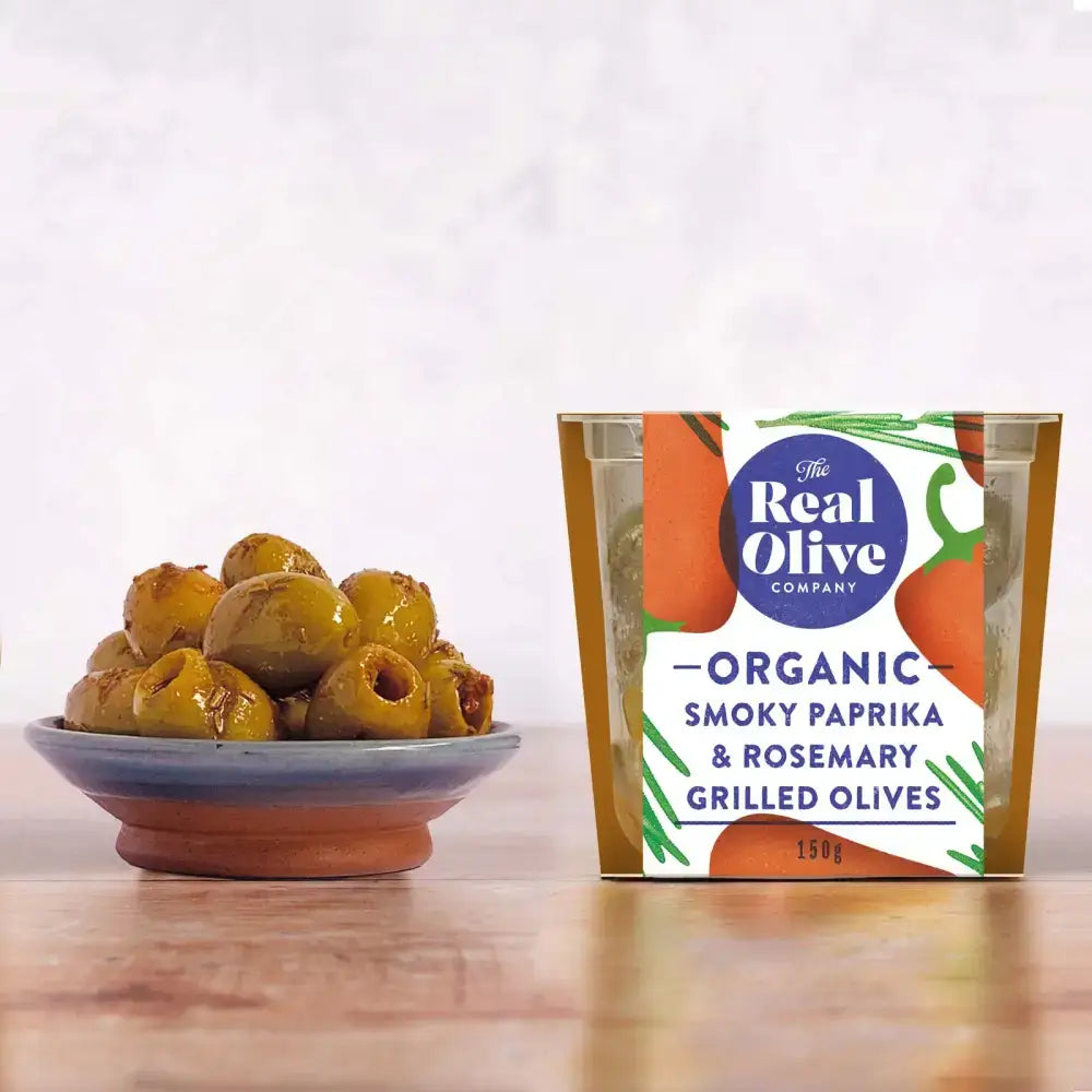 A clear plastic container with a gold lid holds organic smoky paprika and rosemary grilled olives from the real olive company