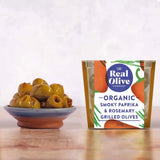 A clear plastic container with a gold lid holds organic smoky paprika and rosemary grilled olives from the real olive company