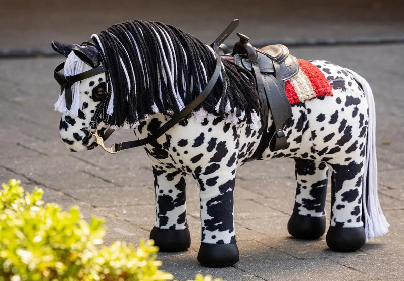 A plush black and white spotted pony toy with a textured mane, fitted with a leather saddle and bridle.