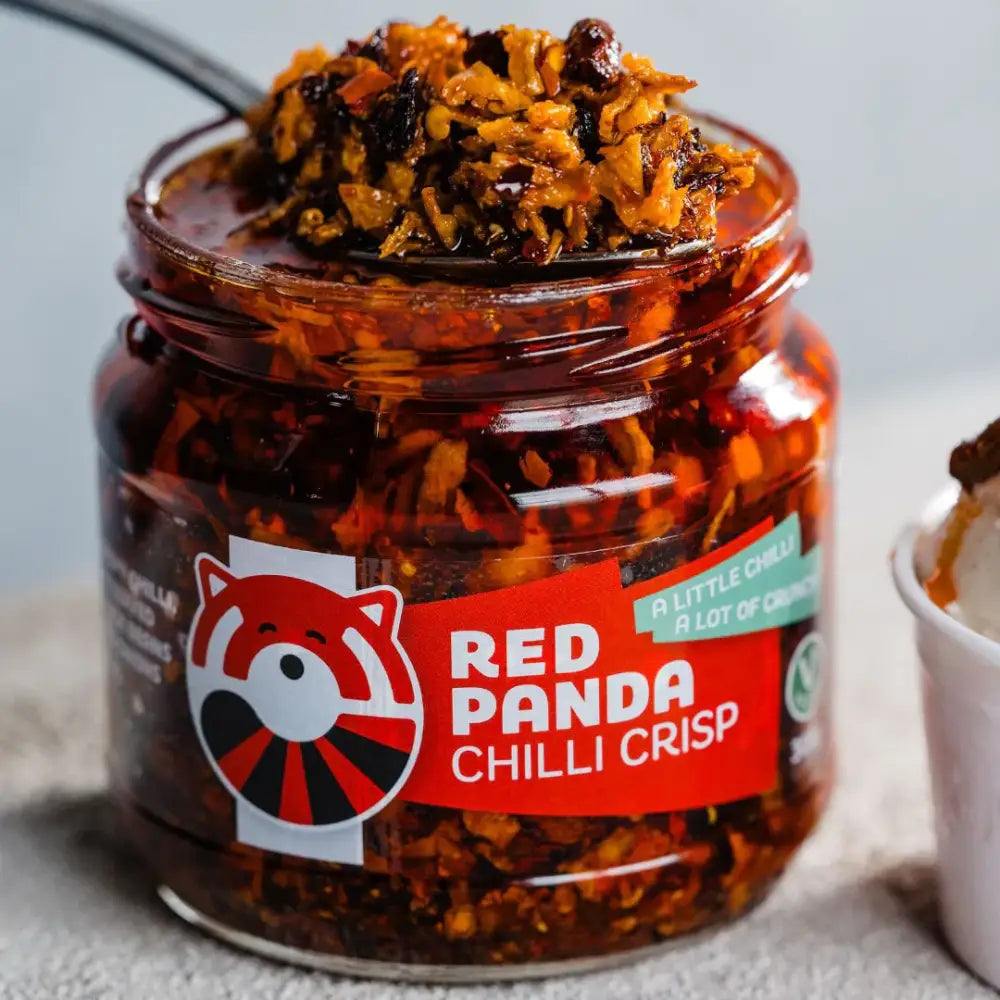 A clear glass jar of red panda chilli crisp filled with vibrant orange-red spicy flakes and a red and black panda logo label