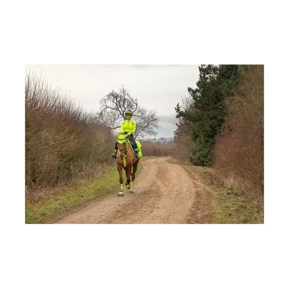 A rider wearing a neon yellow reflector base layer from hy equestrian rides a brown horse along a dirt path