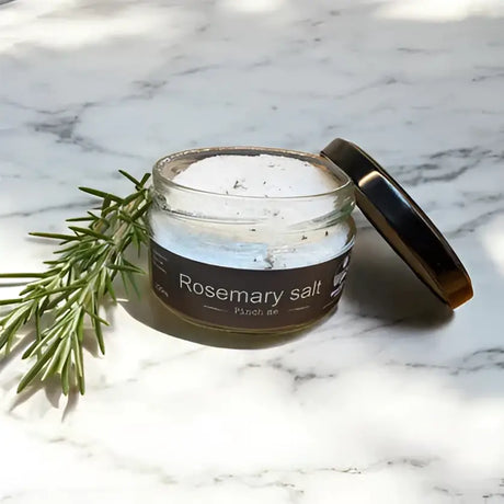 A clear glass jar with a dark brown lid of white rosemary herb infused salt by broomhill farm co labeled rosemary salt