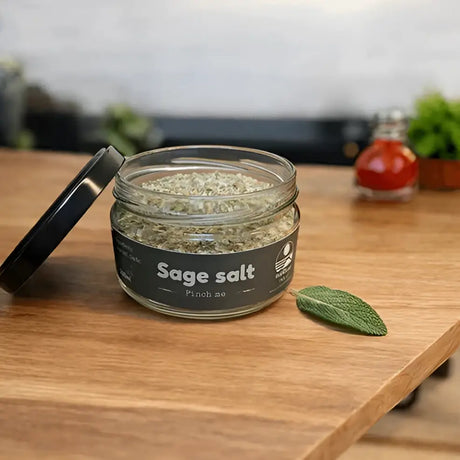 A clear glass jar of greenish sage herb infused salt with a black lid labeled sage salt pinch me by broomhill farm co