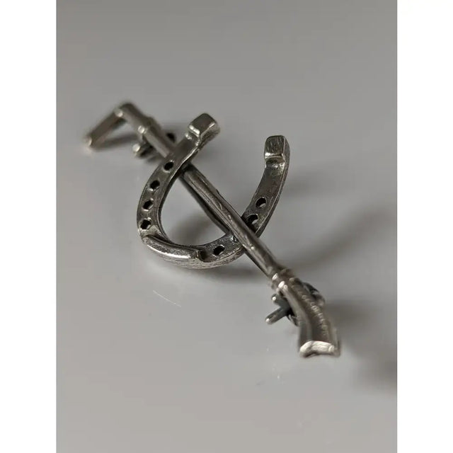 Silver steed shoe stock tie pin featuring a silver metal brooch shaped like a horseshoe crossed with a miniature rifle