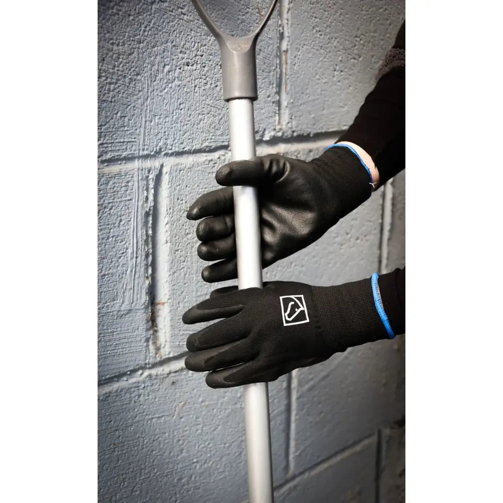 Black ssg equestrian lined barn glove with white logo patch and blue wrist trim gripping a silver tool against gray wall