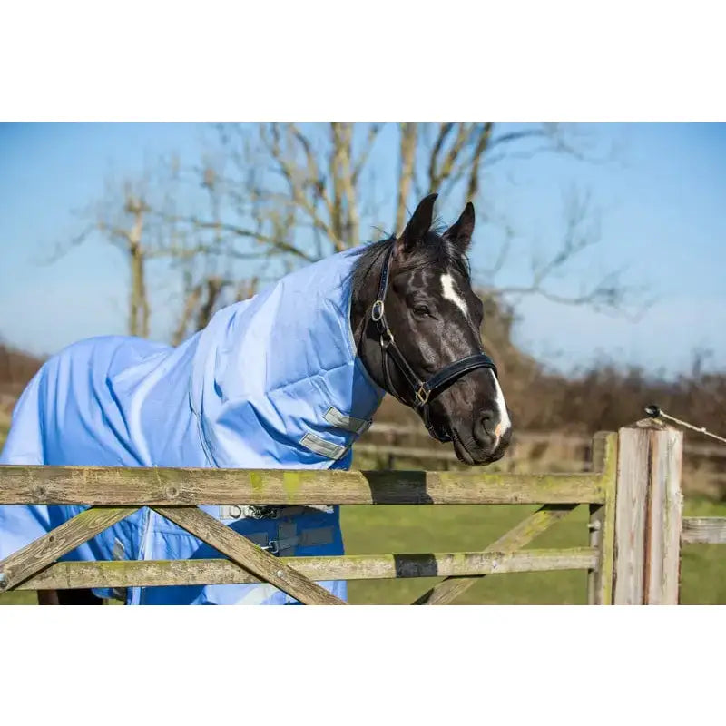 A black horse wearing a cornflower detachable neck turnout rug with 1200d outer stands behind a wooden fence