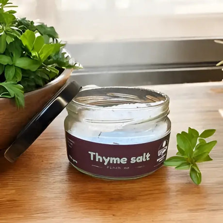 A clear glass jar with black lid labeled thyme salt herb infused salt by broomhill farm co