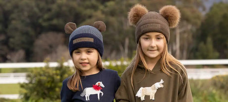 Two children wear knitted beanies with fluffy brown pom-poms and bear ear accents, one in navy blue and the other in olive green.