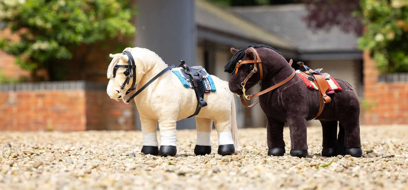 Two plush toy horses, one cream-colored with black hooves and a blue saddle blanket, the other dark brown with tan harnesses and red accents.