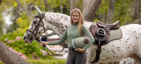 A woman in a sage green long-sleeved top and dark gray riding pants stands beside a white spotted horse, holding its reins.