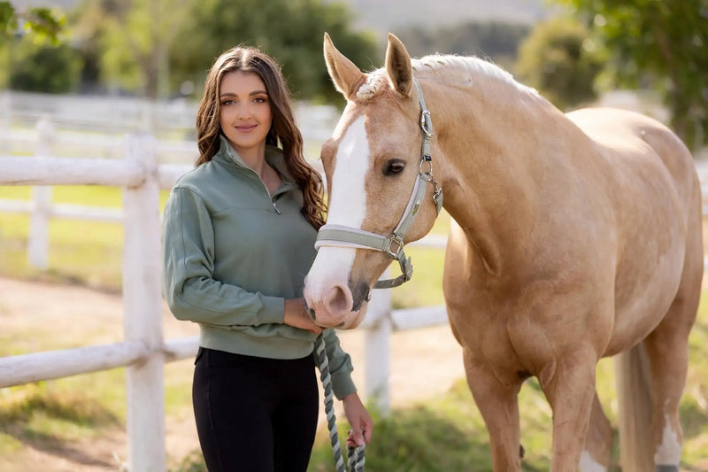 A woman in a sage green quarter-zip pullover stands beside a palomino horse with a white blaze, holding its lead rope.