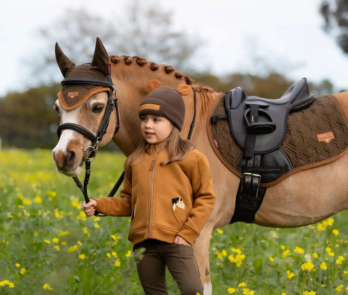 A young girl in a mustard-colored fleece jacket with a horse-themed patch stands beside a light brown horse wearing a black and tan saddle.