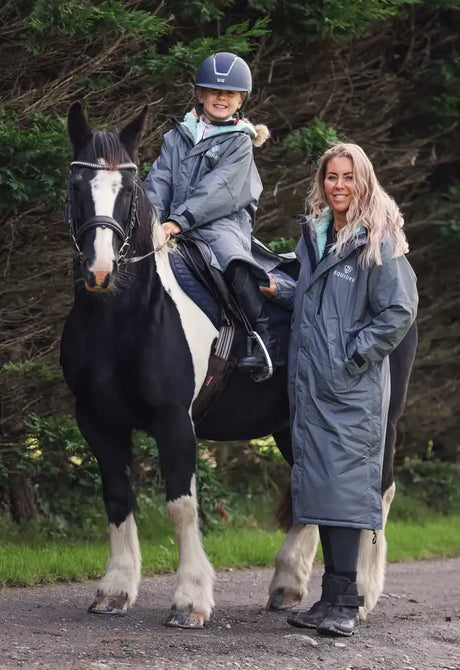 A young girl wearing a navy blue equestrian helmet and a grey riding jacket sits atop a black-and-white horse.
