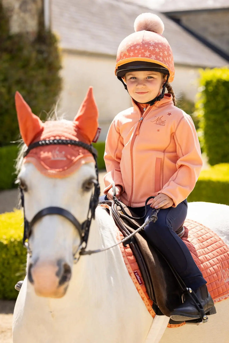 A young girl wearing a pink star-patterned equestrian helmet with a fluffy pom-pom on top.