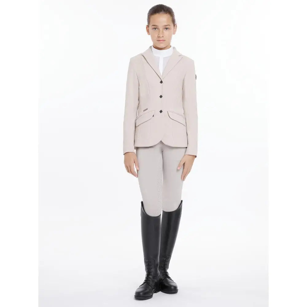 A young girl wearing a lemieux young rider kennedy show jacket stone in beige with black buttons and equestrian boots