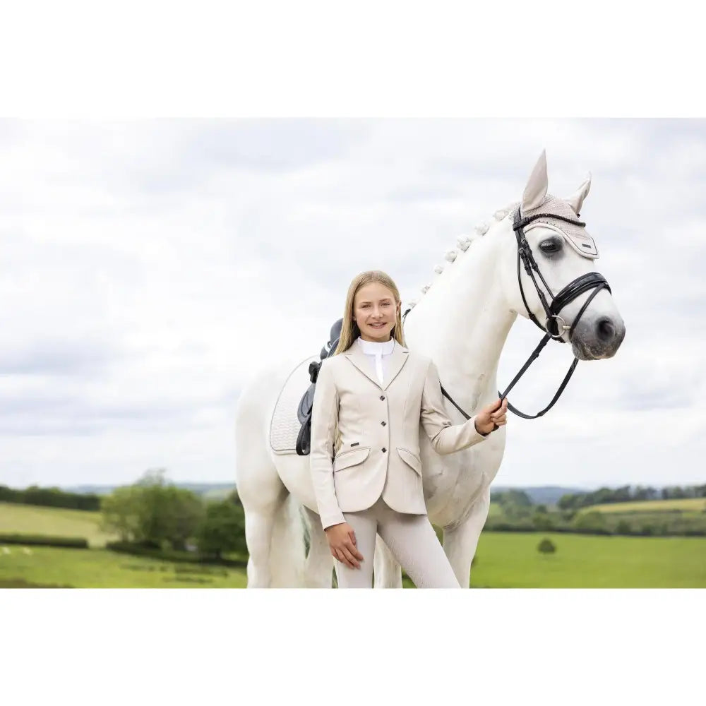 A white horse wearing lemieux black tack and a light hood stands next to a young rider kennedy show jacket stone equestrian gear